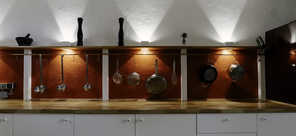 Innenansicht Hofkontor Bonn: illuminierte Küchenarbeitszeile. An einem Kupferrohr hängen Pfannen und Siebe. Auf einer Ablage stehen Salz- und Pfefferstreuer.